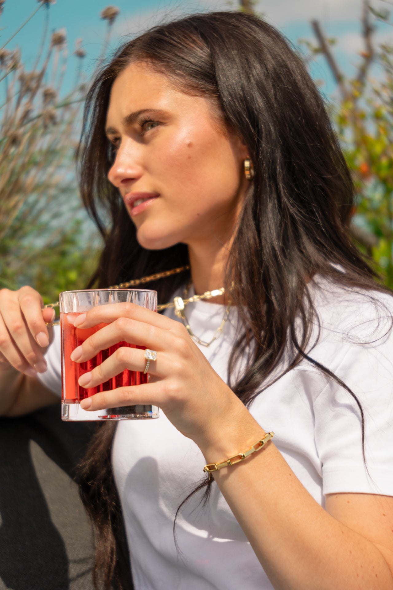 Brunette woman enjoying a refreshing drink outdoors, showcasing her elegant engagement ring. She is wearing a stylish gold bracelet and necklace, which complement her white top. The sunny day and natural background enhance the overall serene and joyful mood of the scene. Jewelry Made by Promise Jewelry 